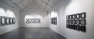 White-walled gallery with a grey floor and track lighting; black-and-white abstract works in square frames, featuring circles and X shapes in rows.