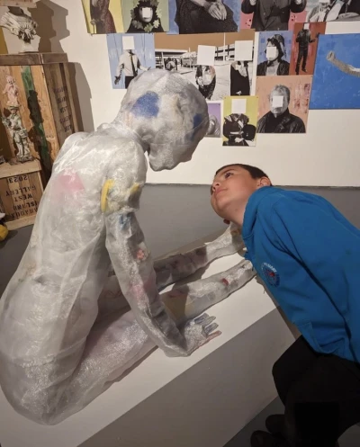 Life-size figure wrapped in bubble wrap sits on a bench, leaning toward a person in a blue hoodie who tilts their head close in an art space.