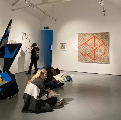 Two people sketch on the gallery floor as a visitor studies a wall piece; right, a large orange cube painting, left, a blue geometric sculpture.