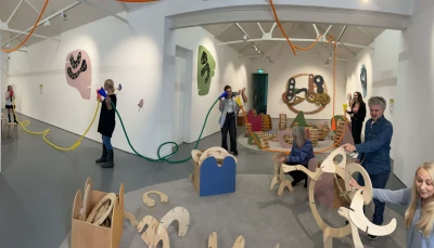 Visitors in a bright gallery interact with a colourful tubular sculpture and wooden curved forms, beside white walls with abstract art.