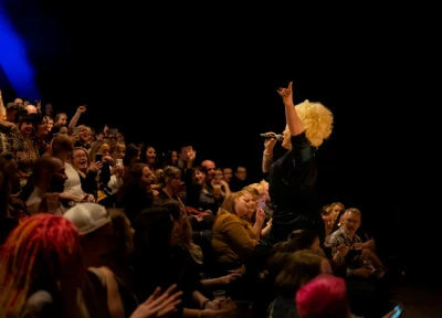 A performer with curly blond hair in a dark, sparkly outfit sings into a microphone on stage, raising an arm while a cheering audience watches.