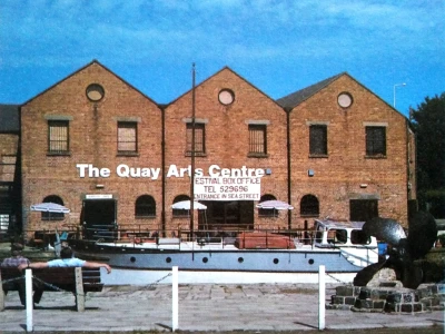 Brick Quay Arts Centre with three gabled sections and circular windows; a moored boat and outdoor seating with umbrellas sit in front of the building.