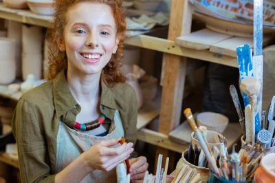 Person with curly red hair smiles in a pottery studio, wearing a green shirt and apron, holding a clay piece, with shelves of brushes and pottery tools behind.