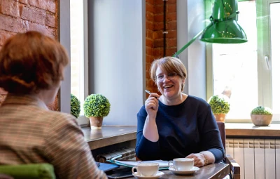Two people chat at a cafe; one faces the camera, smiling and gesturing with a utensil, the other has their back to us. Cups and plants on the sill.