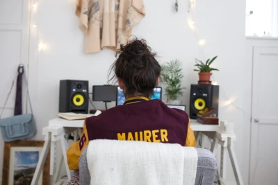 Back view of a person in a burgundy and yellow varsity jacket with 'MAURER' on it, seated at a desk with yellow speakers, plants and string lights.