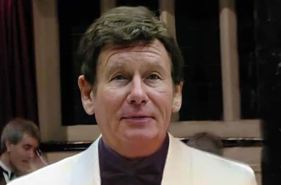 Portrait of a person with short dark hair, wearing a white dinner jacket and dark bow tie, on a stage with a blurred background.