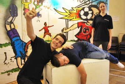 Three people in a gym with a bright mural: one person lying on a white block, another taking a selfie with a phone, and a third standing nearby.
