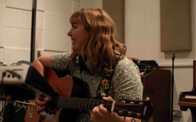 Person with shoulder-length blond hair plays an acoustic guitar in a studio, wearing a green floral shirt and a sunflower-patterned guitar strap.